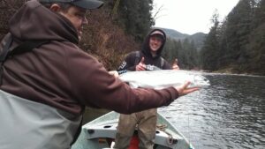 Angler in brown jacket holds silver coho salmon on small boat with forested river banks behind
