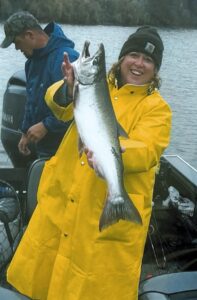 Woman in yellow rain gear holding coho salmon on boat with forested river in background