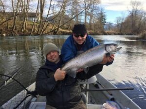 Two anglers in small boat displaying large silver coho salmon on river with bare trees and hillside house