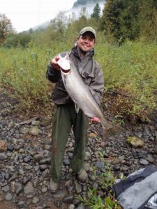 Angler in waders holding large coho salmon on rocky riverbank with misty forested mountains behind
