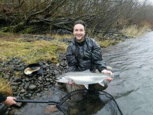 Angler kneeling on rocky riverbank holding large silver coho salmon with fishing net visible.