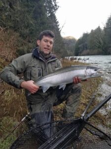 Angler holding large silver coho salmon by river with trees and overcast sky in background