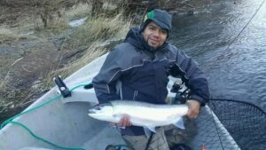 Angler in boat holding fresh coho salmon beside river with grassy bank and flowing water