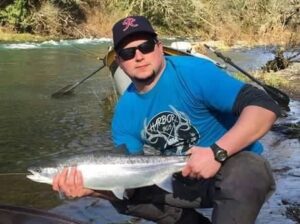 Angler in blue shirt and sunglasses kneeling in shallow river holding silver coho salmon for photo