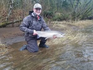 Angler kneeling in shallow water holding a silver coho salmon at muddy riverbank with vegetation.