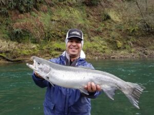 Angler holding large silver coho salmon at riverbank with clear green water and mossy embankment behind