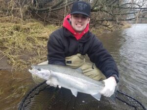 Angler in waders holding large coho salmon while kneeling at muddy riverbank with fallen trees