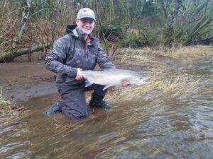 Angler kneeling in shallow water holding silver coho salmon on overcast day at riverbank.