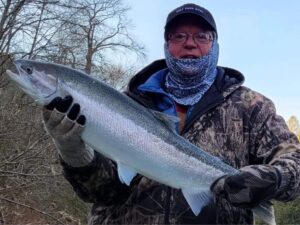 Angler in camouflage holding large silver coho salmon on riverbank with bare trees behind