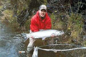 Angler in red jacket holding large coho salmon while standing in shallow river with net