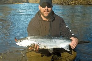Angler in black hoodie and cap holding large silver coho salmon at river's edge with flowing water behind.