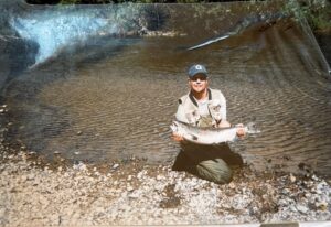 Angler kneeling on rocky riverbank holding silver salmon in shallow water with concrete structure behind