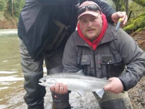 Angler in gray fishing jacket holding fresh coho salmon by river with muddy green water behind