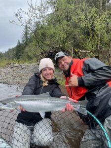 Two anglers pose with a coho salmon in net on rocky riverbank with trees in background.