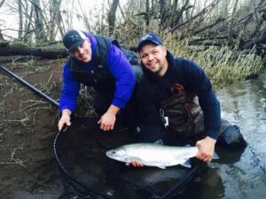 Two anglers kneeling by river holding large silver salmon in landing net on muddy bank.
