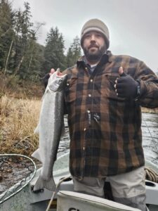 Bearded angler in plaid jacket holding fresh coho salmon in boat on overcast day with forested riverbank.