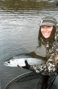 Angler in camouflage holding a bright silver coho salmon over landing net in calm river water.