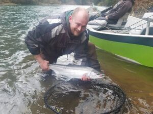 Angler in waders holding coho salmon in shallow water next to green kayak on riverbank