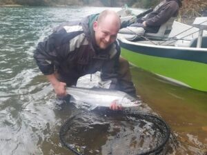 Angler in waders holding coho salmon in shallow water next to drift boat with green hull
