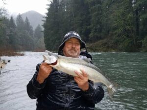 Angler in rain gear holding coho salmon at snowy riverbank with evergreen forest and mountains behind