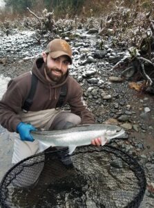 Angler kneeling on snowy rocky riverbank holding coho salmon over landing net in winter conditions