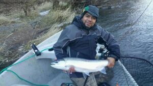 Angler in winter clothing holding a silver coho salmon in small boat on river.