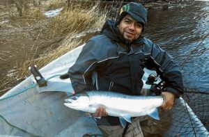 Angler in small boat holding silver coho salmon by river with dry grass banks and snow patches