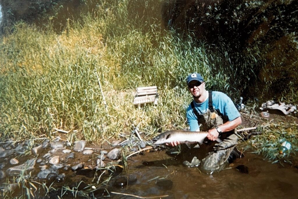 Angler kneeling in shallow river water holding steelhead with tall grass and rocks in background