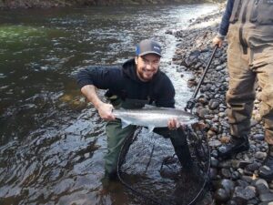 Angler kneeling in shallow river water displaying a steelhead over landing net on rocky shore