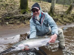 Angler kneeling in shallow river holding large steelhead with moss-covered trees in background