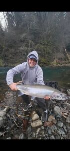 Angler kneeling on rocky riverbank holding large steelhead with dog beside him, forested background.