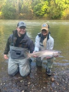 Two anglers kneeling in river holding large steelhead trout with forested riverbank behind them