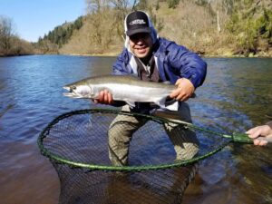 Angler in blue jacket holding steelhead over landing net in clear river with forested hillside background.