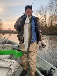 Angler holding large steelhead at sunset beside kayak on river bank with bare trees in background