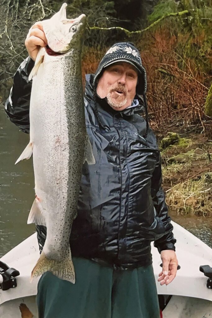 Angler in camouflage jacket holding large steelhead while standing in boat on forested river