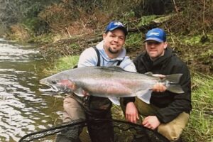 Two anglers kneeling by river holding large steelhead trout for photo on grassy bank