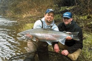 Two anglers kneeling by river holding large steelhead trout for photo on grassy bank