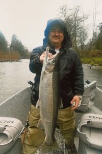 Angler in rain gear holding large steelhead in aluminum drift boat on overcast river day