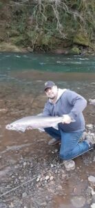 Angler kneeling on rocky riverbank holding large steelhead beside flowing green water with overhanging vegetation