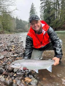 Angler in red and black jacket kneeling with steelhead on rocky riverbank with evergreen forest background