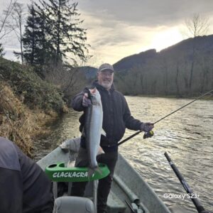 Angler in drift boat holding large steelhead on overcast day with forested hillside in background