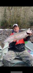 Angler sitting on riverbank holding large steelhead with bare deciduous trees and rocky shore in background