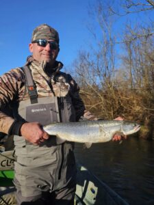 Angler in camouflage and waders holding steelhead in drift boat on clear winter day