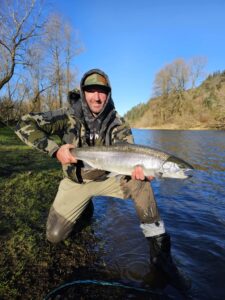 Angler kneeling in shallow river water holding large steelhead with forested hillside and blue sky in background.