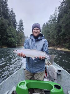 Angler holding steelhead in small boat on forested river in overcast conditions wearing beanie and rain jacket