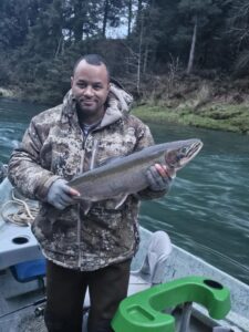 Angler in camouflage jacket holding steelhead on boat in forested river setting with green water
