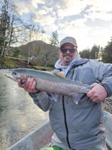 Angler in gray jacket holding large steelhead trout at riverbank with trees and hills in background