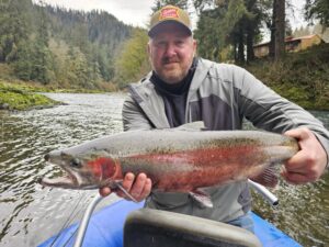 Angler in boat holding large steelhead trout on forested river with cabin visible in background
