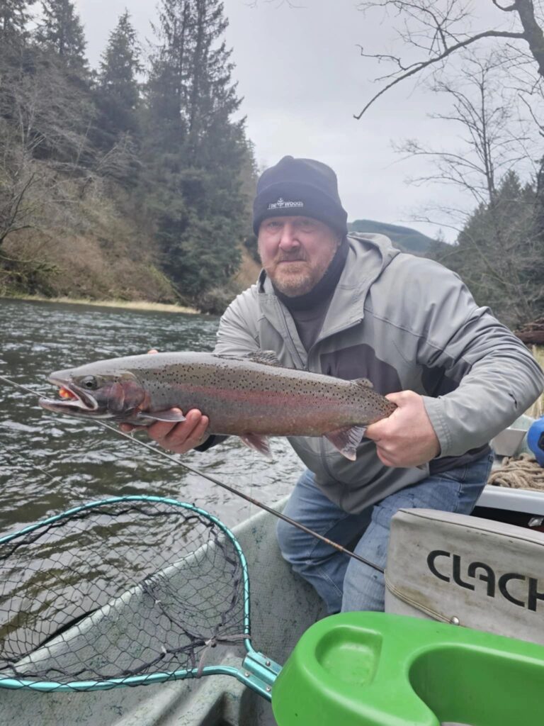 Angler holding large steelhead trout in boat on misty river surrounded by evergreen forest