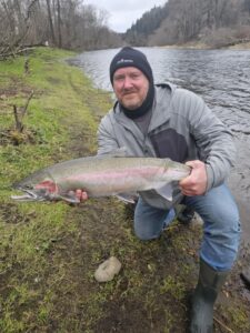Angler kneeling on grassy riverbank holding large steelhead with distinctive pink stripe along lateral line.